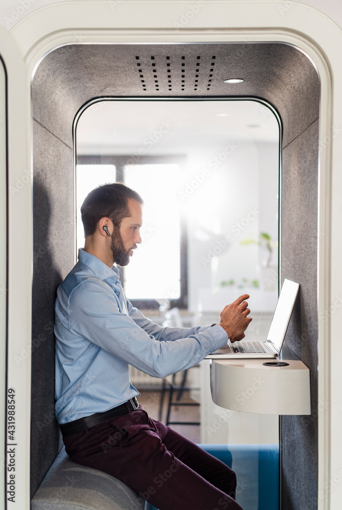 Male entrepreneur attending meeting on laptop in telephone booth at office Male entrepreneur attending meeting on laptop in telephone booth at office