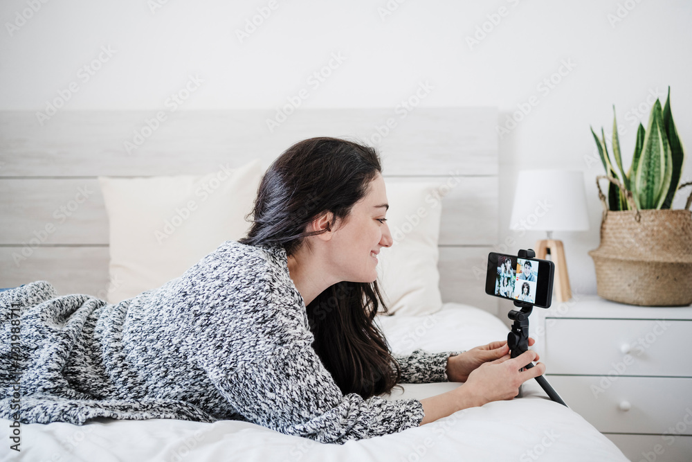 Smiling woman talking on video conference through mobile phone while lying on bed at home