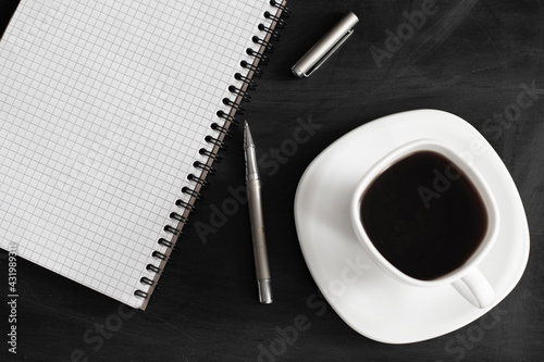 White cup of strong black coffee with metal pen and notepad on black surface close up