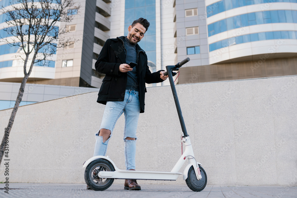 Smiling handsome man using smart phone while standing by electric push scooter against buildings Smiling handsome man using smart phone while standing by electric push scooter against buildings