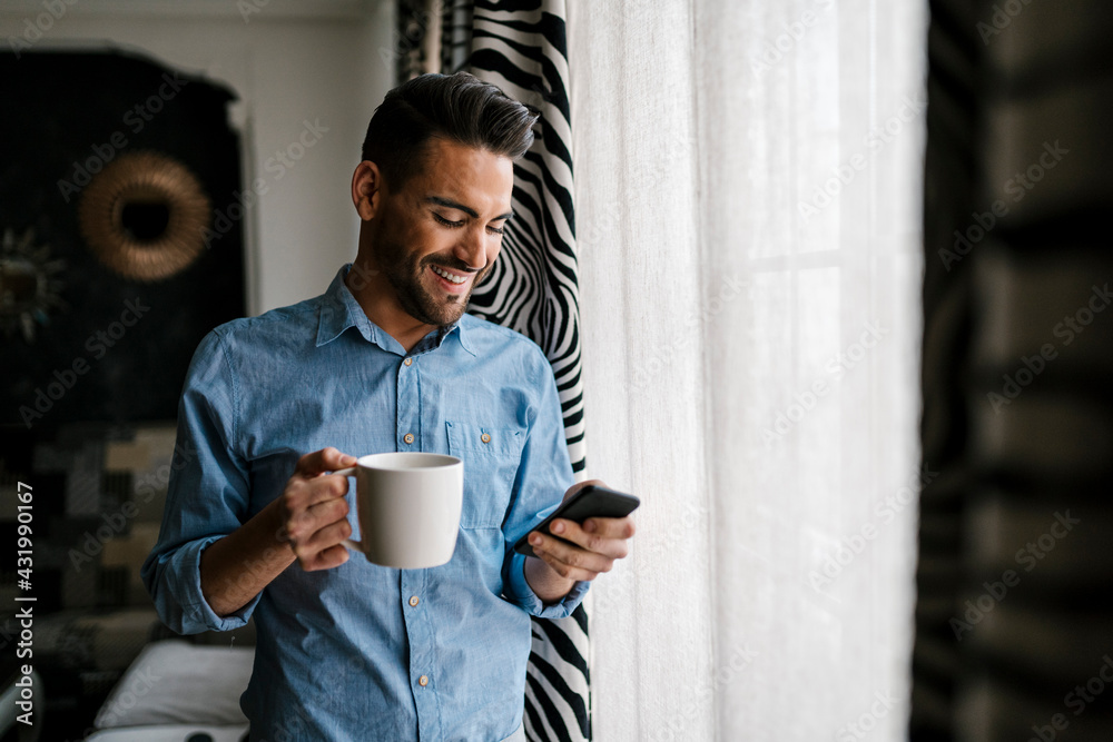 Smiling mid adult man with coffee cup using mobile phone while standing at home Smiling mid adult man with coffee cup using mobile phone while standing at home