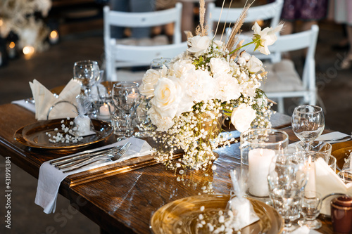 Wedding decor of roses, cotton and gypsophila and candles on a laid festive table with glasses and plates with cards. Off-season rustic wedding dinner. Selected focus and blurred background.