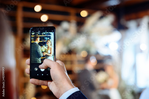 Wedding ceremony of the groom in a tuxedo and the bride in a white dress. The couple looks at each other against the backdrop of the wedding arch. The guest is filming what is happening on the phone