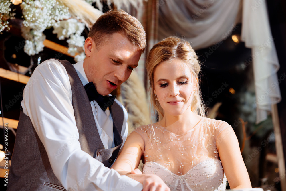 Naklejka premium A groom in a tuxedo and a bride in a dress cut the wedding cake at the end of the evening. Wedding traditions of the couple. Emotions and joy from starting a family.