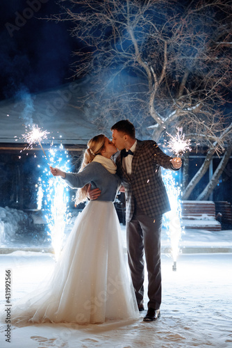 A groom in a jacket and a bride in a jacket are holding sparklers and fireworks in their hands. Kisses against the background of cold sparkling fountains outside in winter. 