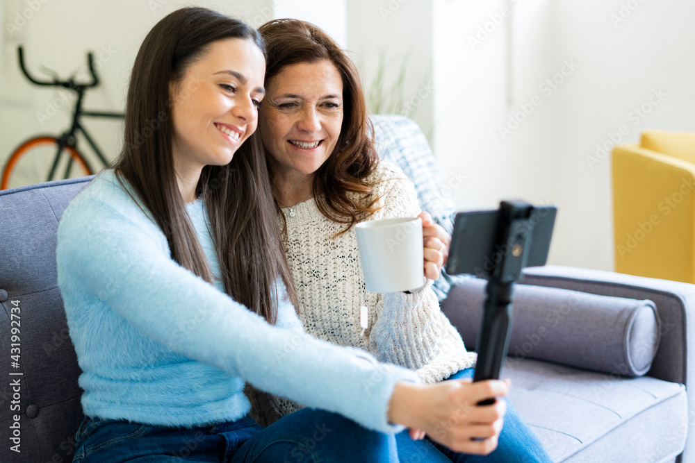 Smiling daughter and mother vlogging through smart phone while sitting on sofa at home Smiling daughter and mother vlogging through smart phone while sitting on sofa at home