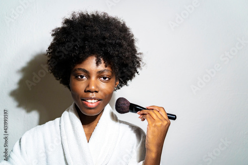 Curly woman using blushbrush while standing against white background photo