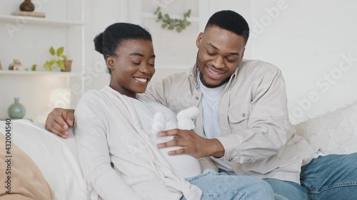 African American couple family future expecting parents sitting on couch afro black husband man stroking wife pregnant belly with cute small childish child white shoes hugging embracing beloved woman