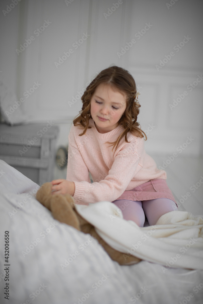 A beautiful little girl with curly hair stands at the window in a bright room and puts a teddy bear to sleep on a white bed A beautiful little girl with curly hair stands at the window in a bright room and puts a teddy bear to sleep on a white bed