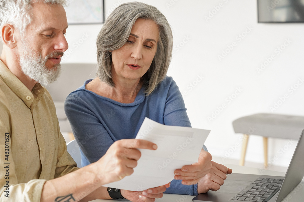 Older mature couple checking bank documents using laptop at home. Senior mid age retired man and woman reading paper bills, calculating pension or taxes, planning retirement finances, doing paperwork. Older mature couple checking bank documents using laptop at home. Senior mid age retired man and woman reading paper bills, calculating pension or taxes, planning retirement finances, doing paperwork.