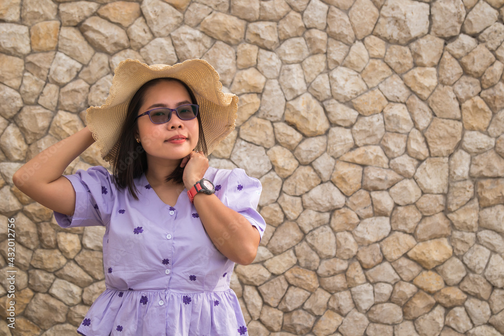 Asian woman dressed in purple shirt wearing glasses and straw hat on brick wall background.