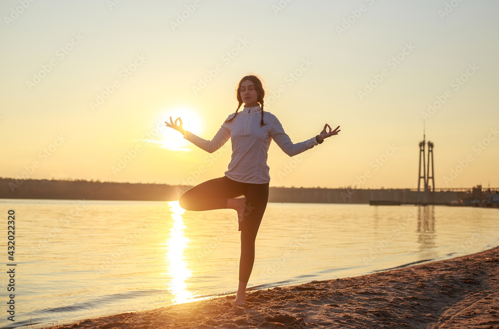 Fototapeta premium Young woman meditating doing yoga on sandy beach by the river on background of setting sun