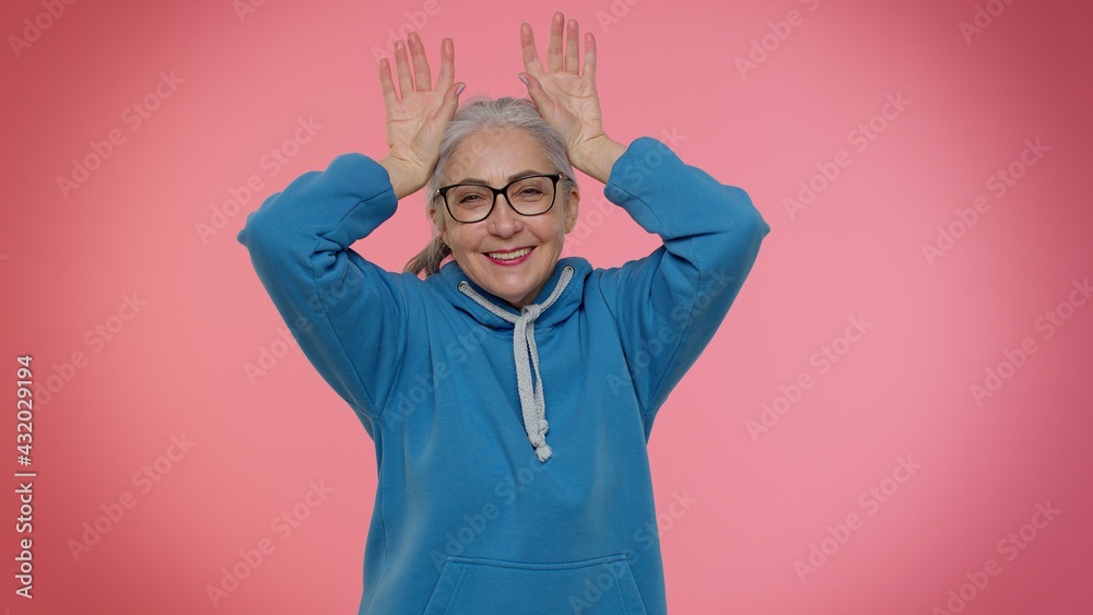 I am little bunny rabbit. Funny mature old granny woman smiling friendly and doing bunny ears gesture on head, having fun, fooling with humorous comical mood. Senior grandmother on pink background