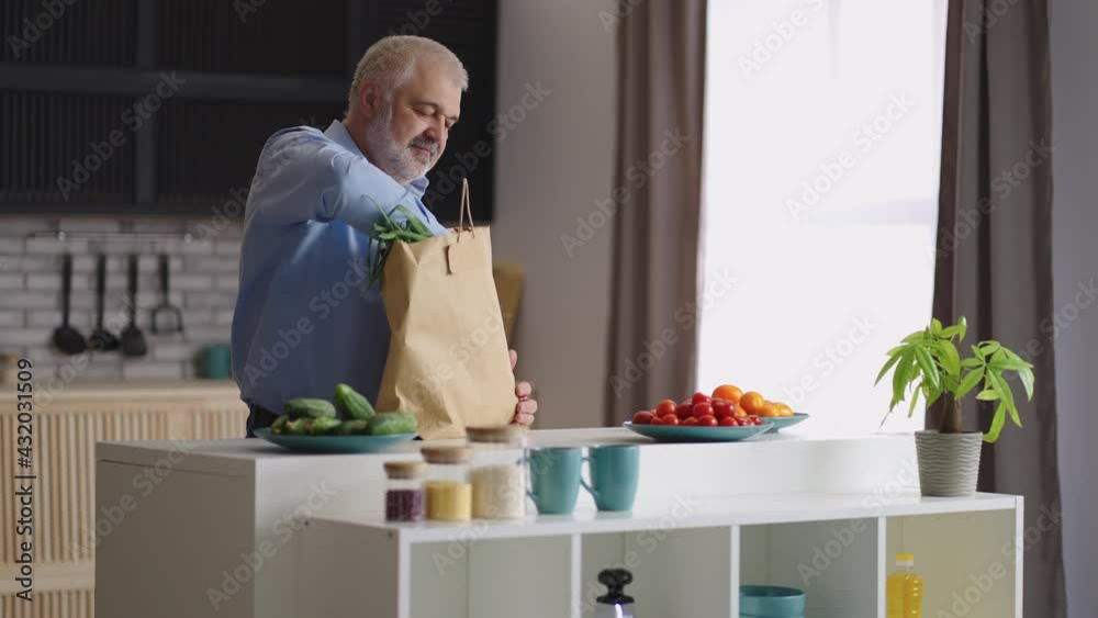 grey haired man is unpacking food after shopping in grocery, putting fresh greenery and baguette on kitchen table grey haired man is unpacking food after shopping in grocery, putting fresh greenery and baguette on kitchen table