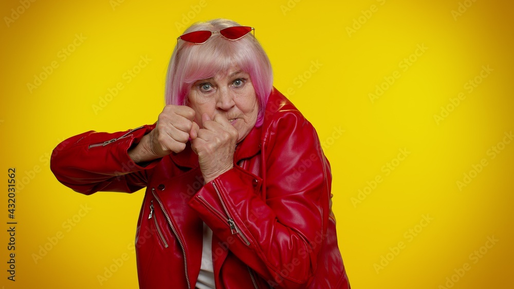 Funny elderly rocker granny woman trying to fight at camera, shaking fist, boxing with expression, punishment. Senior stylish old grandmother isolated on yellow background. People lifestyle emotions