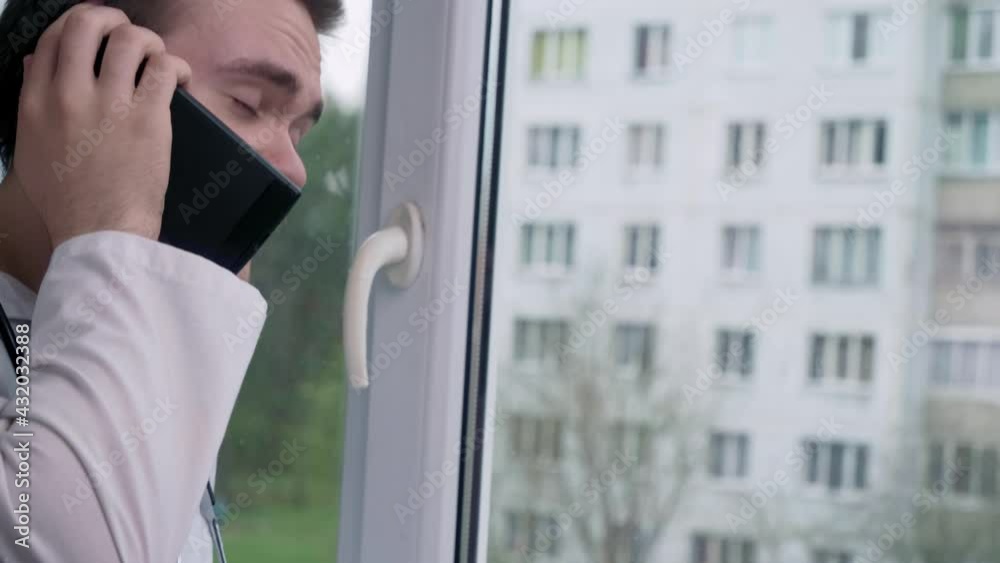 Young doctor speaks on the phone, near the window, against the background of a building, the concept of therapy and a doctor Young doctor speaks on the phone, near the window, against the background of a building, the concept of therapy and a doctor