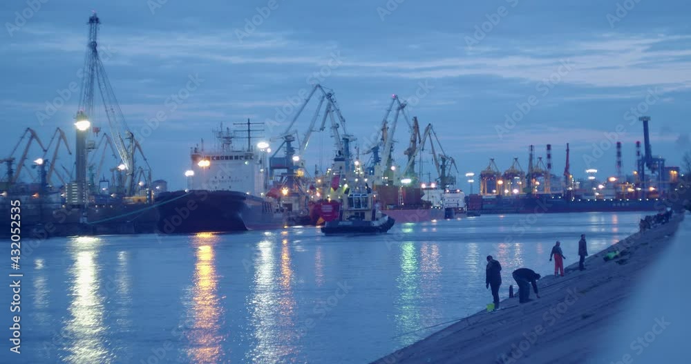 port in the evening, the ship sails along the pier with merchant ships and various cranes, the lights are blurred in the water, fishermen are fishing.