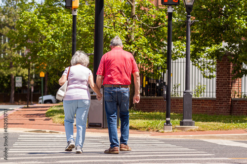 Senior caucasian couple in love holding hands as they cross the street on pedestrian crossing on a lovely spring day. Activity, togetherness at elderly community concept. Woman carries a sidebag.