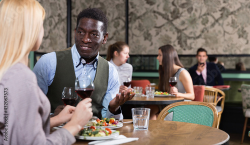 Confident African American businessman with happy cheerful smiling female partner having dinner and drinking red wine at restaurant
