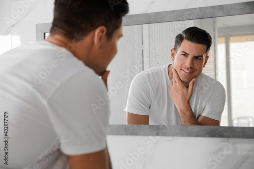 Handsome man touching his smooth face after shaving near mirror in bathroom