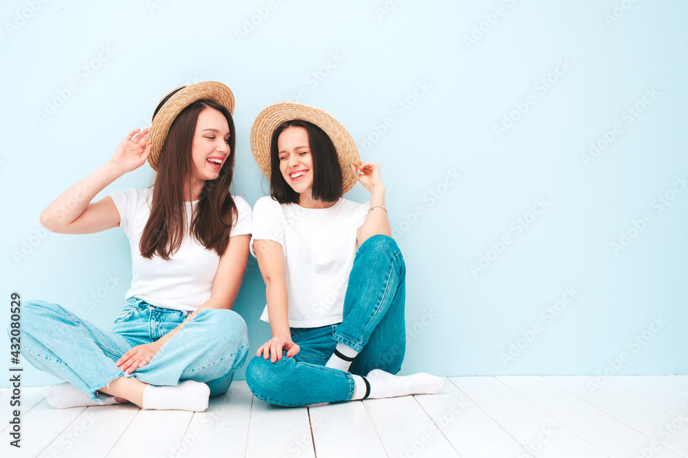 Two young beautiful smiling hipster female in trendy summer white t-shirt and jeans clothes.Sexy carefree women posing near light blue wall in studio.Positive models in hats
