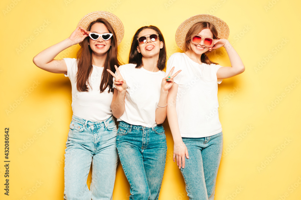 Three young beautiful smiling hipster female in trendy summer white t-shirt and jeans clothes.Sexy carefree women posing near yellow wall in studio.Positive models in hats show peace sign