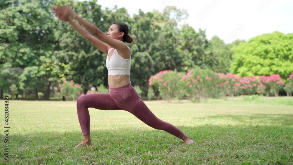 Asian young woman standing on the grass practicing yoga in the Warrior Pose position in city park with the big trees background. Rearview of females practicing yoga outdoors on a sunny day. Asian young woman standing on the grass practicing yoga in the Warrior Pose position in city park with the big trees background. Rearview of females practicing yoga outdoors on a sunny day.