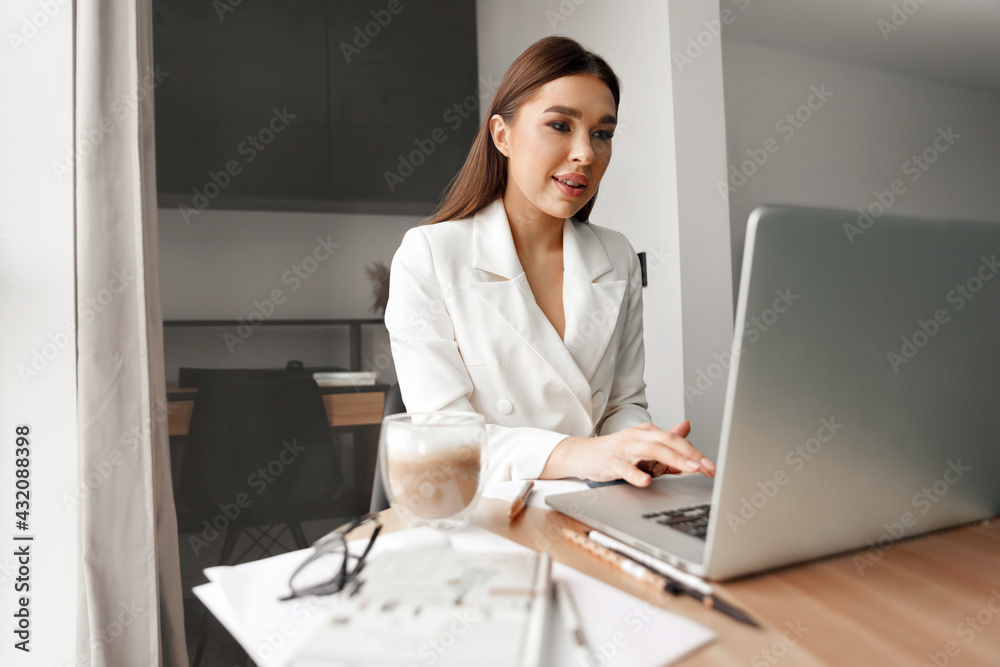 Young female in glasses using laptop, communicates on internet with customer in home, coffee mug on table.Cozy office workplace, remote work, Elearning concept Young female in glasses using laptop, communicates on internet with customer in home, coffee mug on table.Cozy office workplace, remote work, Elearning concept