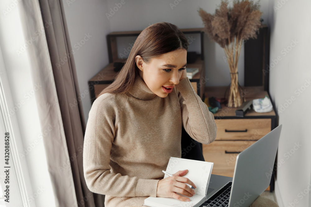 Young beautiful woman working in the home uses a laptop and mobile phone. Female writing in block note Young beautiful woman working in the home uses a laptop and mobile phone. Female writing in block note