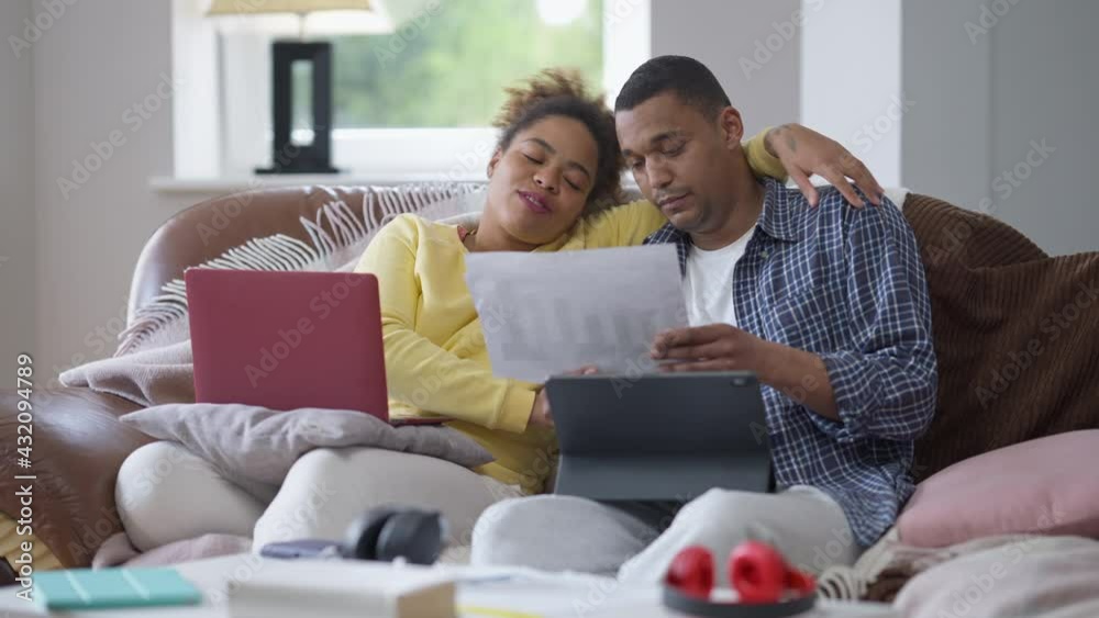 African American man and woman analyzing graphs talking and smiling sitting in home office on couch. Positive couple working from home on coronavirus pandemic quarantine. Family and business African American man and woman analyzing graphs talking and smiling sitting in home office on couch. Positive couple working from home on coronavirus pandemic quarantine. Family and business