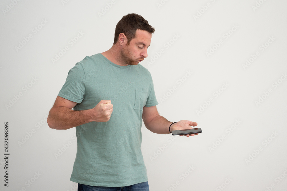 Portrait of a furious man looking at his smartphone, which he holds in one hand and points with the closed fist of the other hand. 