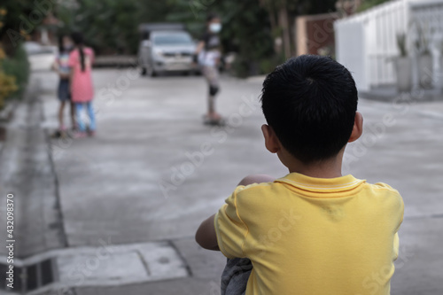 Rear side of black hair young boy wearing yellow shirt sitting hugging knees on cement ground and looking his friends playing skateboard. Selective focus.