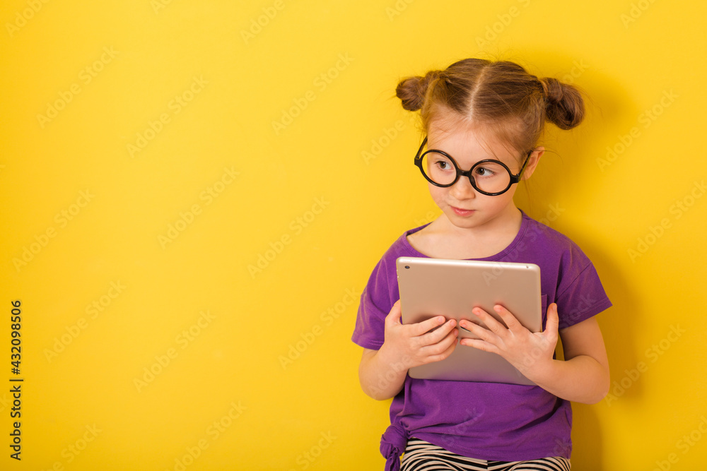 Little cute caucasian funny kid child baby girl with tablet in purple T-shirt on yellow wall background. children studio portrait of student Little cute caucasian funny kid child baby girl with tablet in purple T-shirt on yellow wall background. children studio portrait of student