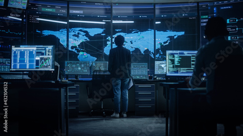 Diverse Multiethnic IT Technical Support Manager with Curved Hair Stands next to Workplace in Monitoring Control Room Office with Big Digital Screens with Server Technical Blockchain Data.