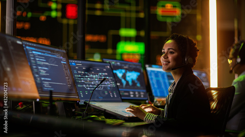 Multiethnic Female IT Technical Support Specialist Talking with a Client in Headphones and Working on Desktop Computer in Monitoring Control Room with Big Digital Screens with Server Blockchain Data.