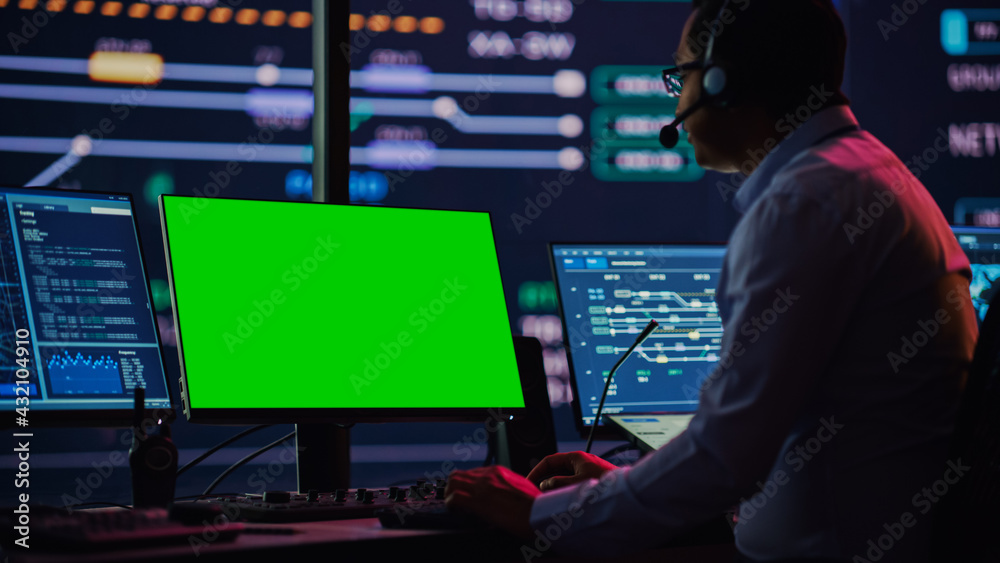 Professional IT Technical Support Specialist and Software Developer Working on Computer with Green Screen Mock Up Display in Monitoring Control Room with Digital Screens. Employee Uses on Headphones. Professional IT Technical Support Specialist and Software Developer Working on Computer with Green Screen Mock Up Display in Monitoring Control Room with Digital Screens. Employee Uses on Headphones.