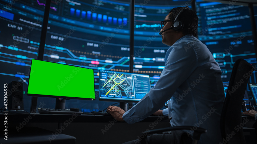Professional IT Technical Support Specialist and Software Developer Working on Computer with Green Screen Mock Up Display in Monitoring Control Room with Digital Screens. Low Angle Shot. Professional IT Technical Support Specialist and Software Developer Working on Computer with Green Screen Mock Up Display in Monitoring Control Room with Digital Screens. Low Angle Shot.