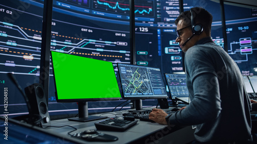 Professional IT Technical Support Specialist and Software Developer Working on Computer with Green Screen Mock Up Display in Monitoring Control Room with Digital Screens. Employee Wears Headphones.