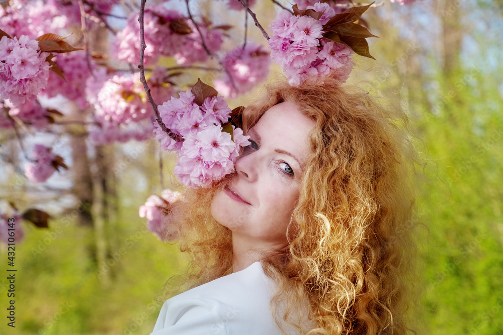 Naklejka premium sensual portrait of a sexy, smiling mature redhead woman in white dress in pink flower tree blossoms in april, spring awakening