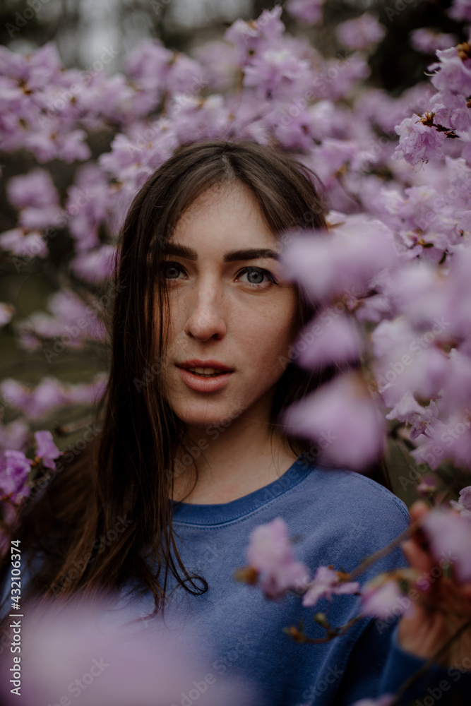 Fototapeta premium young beautiful woman look forward with her mouth covered. portrait of a beautiful woman in the garden near a blossoming tree and its pink branches. brunette girl trying to say something