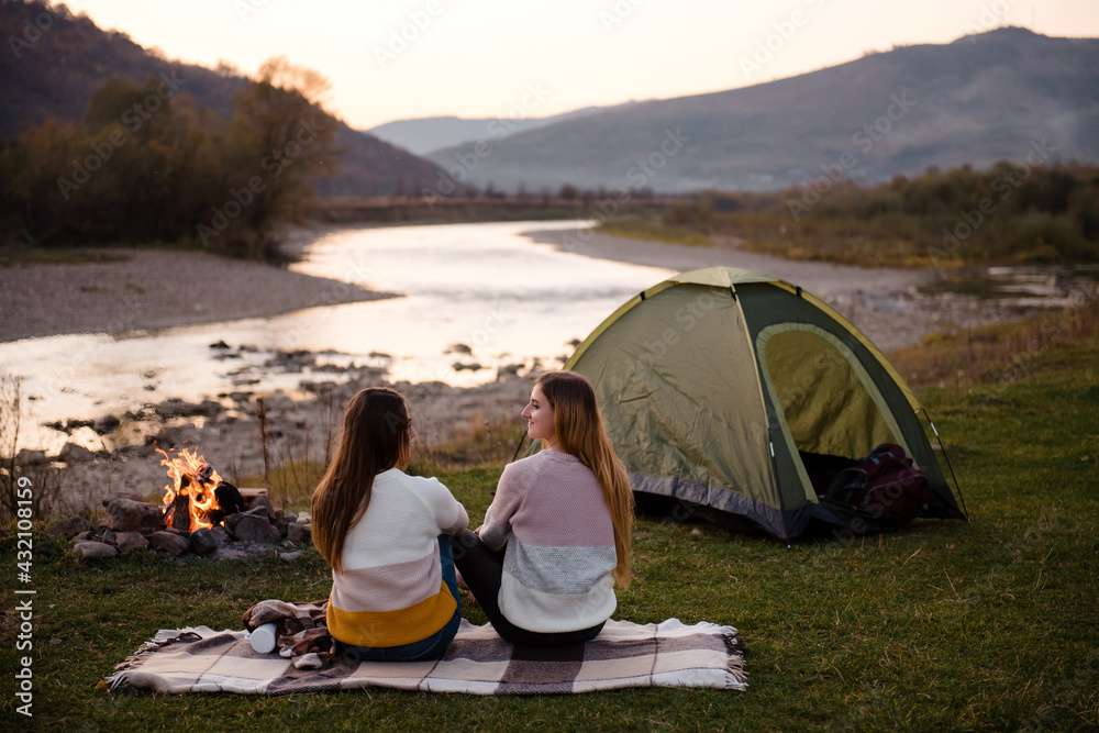 Rear view of two women near tent and campfire. Friends are sitting on the grass on a blanket. Enjoy the scenery of mountains and rivers. Hiking, vacation and friendship concept.