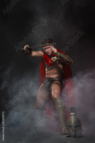 Young athletic sexy man in a costume of a Roman warrior in a red cloak in move on a black background in the smoke.