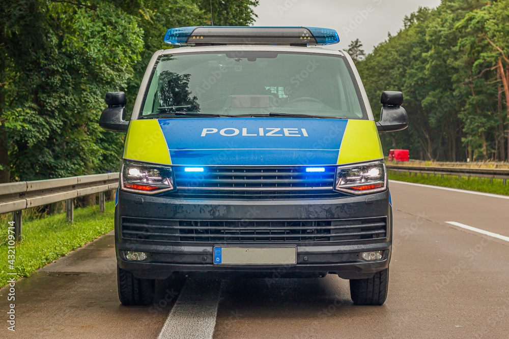 Front view of a police car on the highway. Two-lane motorway in rainy weather. Activated blue light and headlights in the front area. Guard rails and trees in the background