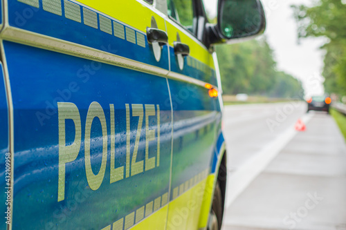 View past a German police car on the autobahn. Lettering on the body with reflective stripes on a blue and yellow background. Rainy lanes and trees. Vehicle in the background on the hard shoulder