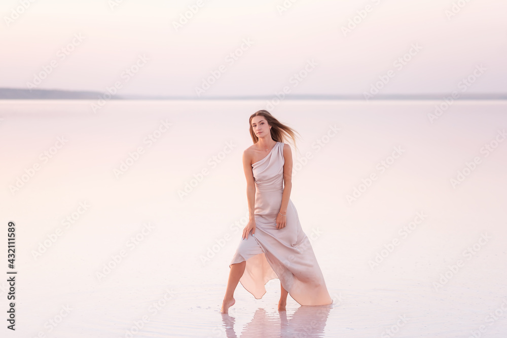 Young blonde woman in an evening airy pastel pink, powdery dress stands barefoot on white crystallized salt. Girl with natural make-up, hair is developing. Salt mining trip, walking on water at sunset Young blonde woman in an evening airy pastel pink, powdery dress stands barefoot on white crystallized salt. Girl with natural make-up, hair is developing. Salt mining trip, walking on water at sunset