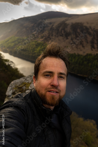 A Young man with beard taking a selfie in mountain with a lake in the background in Wicklow ireland