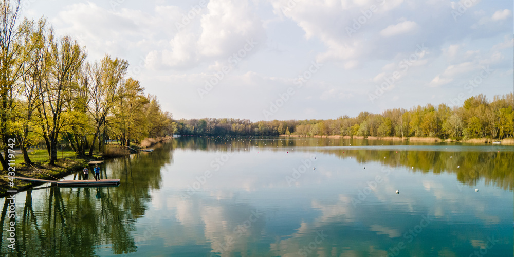 Fototapeta premium Two unidentifiable people with mask and mobile covering faces, standing Lake for canoeing around Danube river in autumn spring colors with blue sky above and reflection in the lake, Zemnik, Slovakia