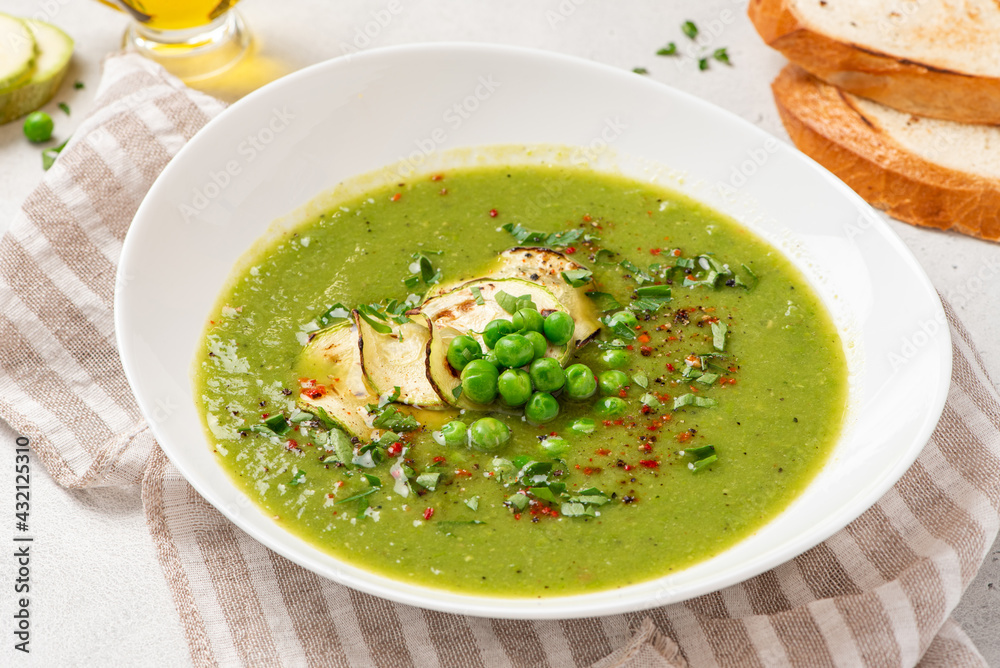 Green cream soup in a white bowl on a concrete background. Vegan cream soup from fresh green peas and zucchini. Tasty diet soup. Green cream soup in a white bowl on a concrete background. Vegan cream soup from fresh green peas and zucchini. Tasty diet soup.