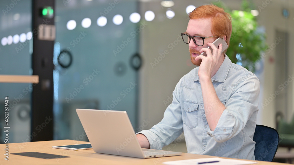 Young Redhead Man with Laptop Talking on Phone, Negotiation in Office Young Redhead Man with Laptop Talking on Phone, Negotiation in Office