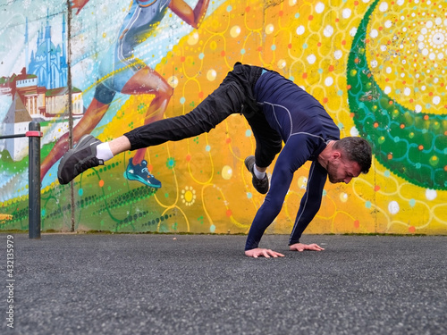 A guy in a navy longsleeve and black pants sits on a street platform doing a handstand. There is a bright wall in the background.